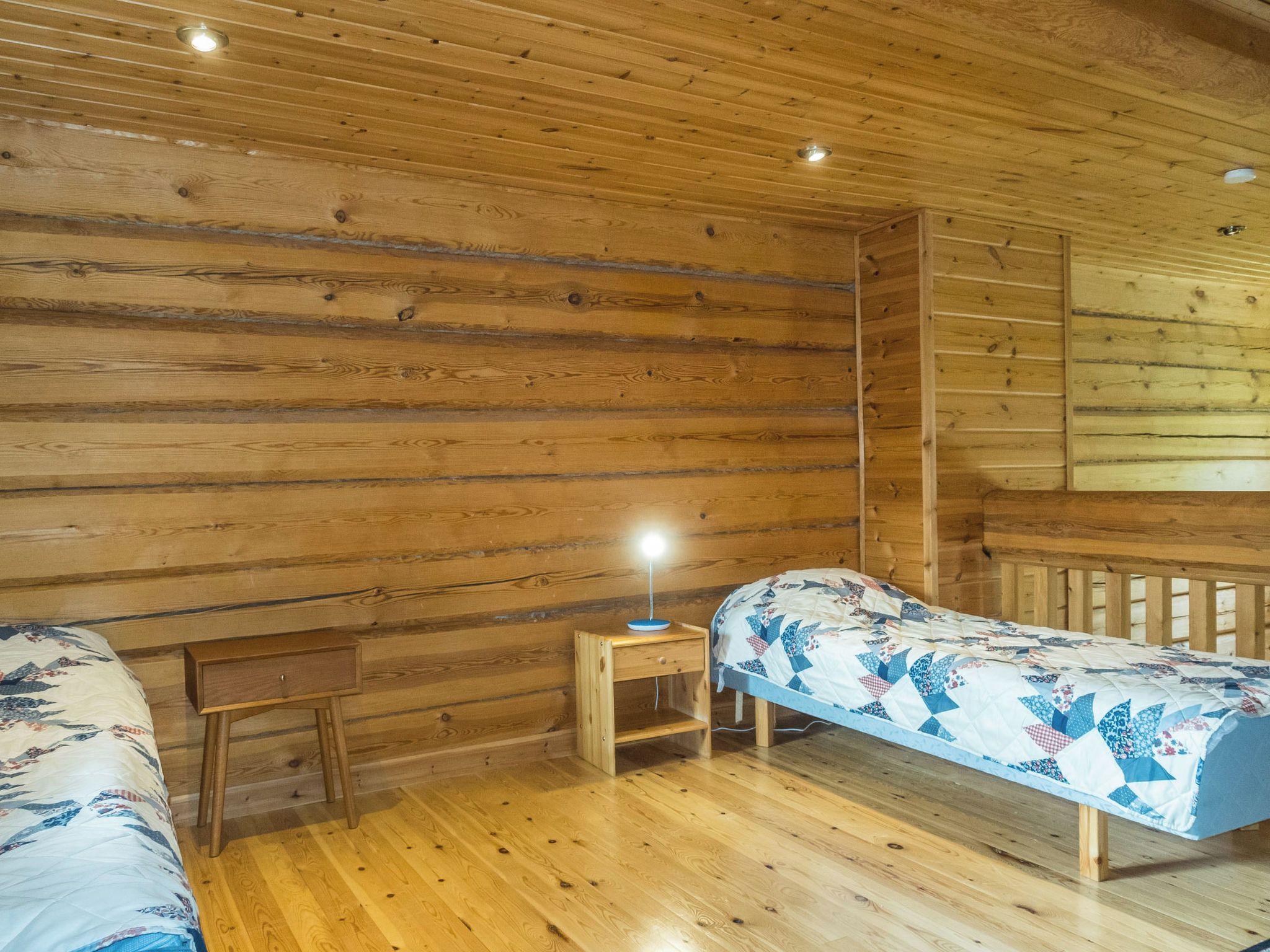 A cabin-style room with wooden walls and floors, two single beds with floral bedding, a small wooden nightstand with a lamp, and a wooden bench.