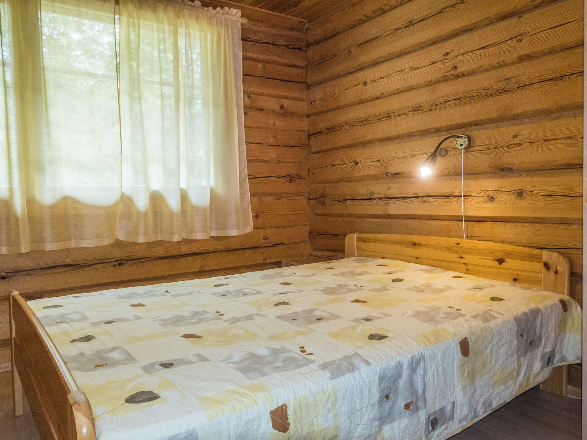 Ein Einzelbett mit einem gemusterten Überwurf in einer Holzhütte mit Holzwänden und einem Fenster mit durchsichtigen Vorhängen.
