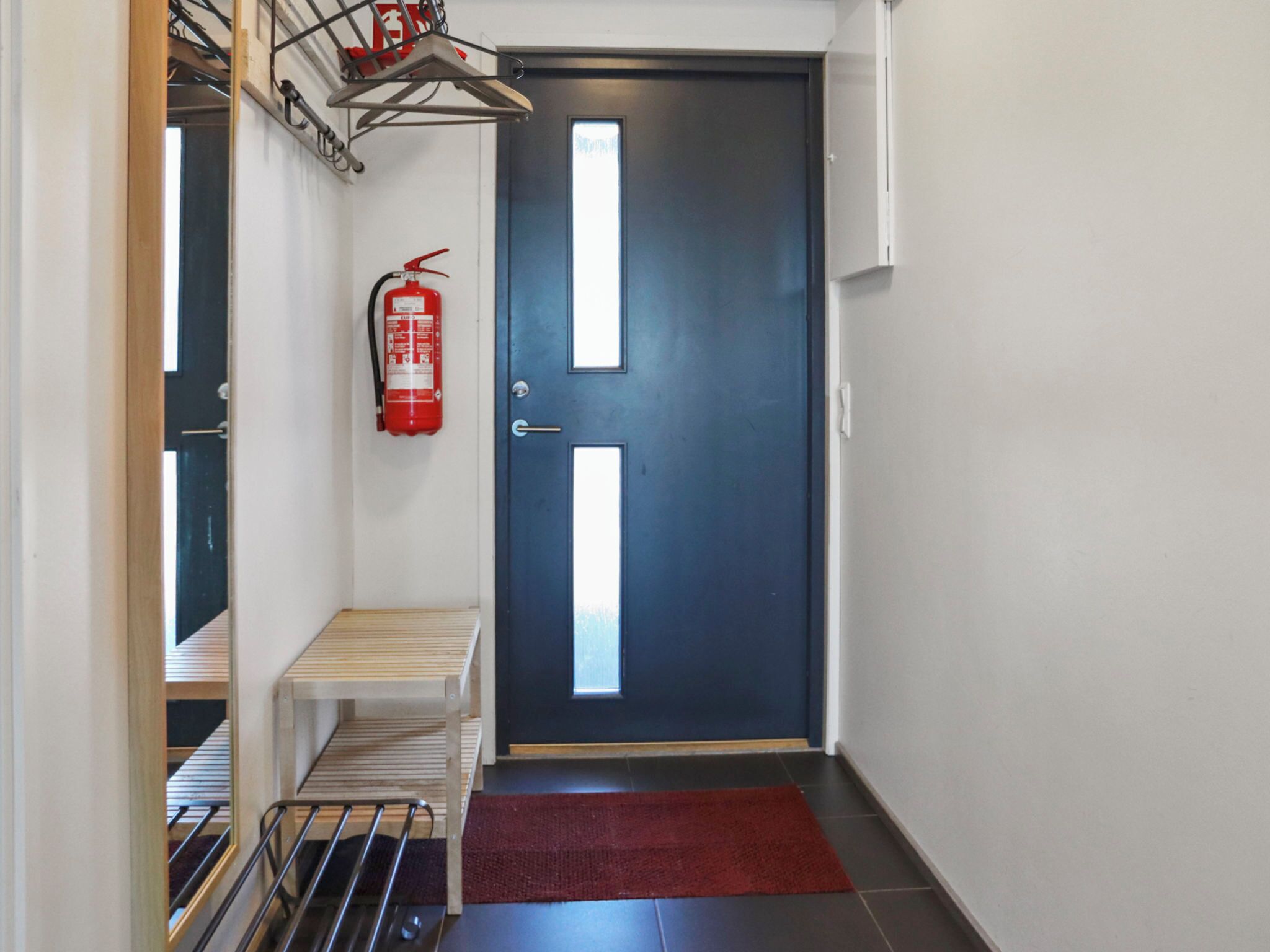 A hallway with a blue door, a fire extinguisher, a small bench, and a red rug.