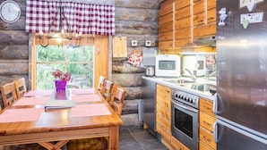 A rustic cabin kitchen with a wooden table, chairs, a refrigerator, a stove, and a window with a view of greenery.