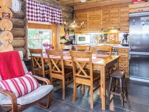 A cozy cabin-style kitchen with wooden cabinetry, a dining table with chairs, a refrigerator, and a window with a view of greenery.
