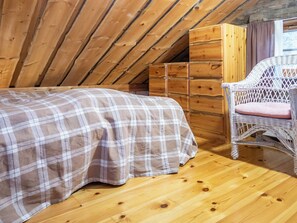 A bedroom with a wooden ceiling, a bed with a plaid bedspread, a wicker chair with a cushion, and a wooden dresser.