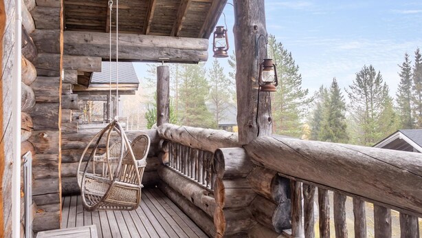 A wooden log cabin with a porch featuring a hanging chair, lanterns, and a view of trees.