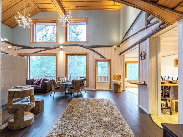 A spacious living area with a high ceiling, wooden beams, and large windows offering views of the outdoors.