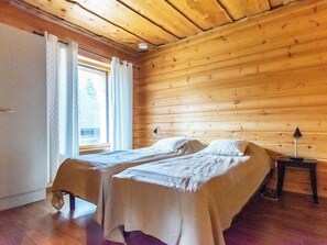 Una habitació amb dos llits, parets de fusta, una finestra amb cortines i una tauleta de nit amb una làmpada.