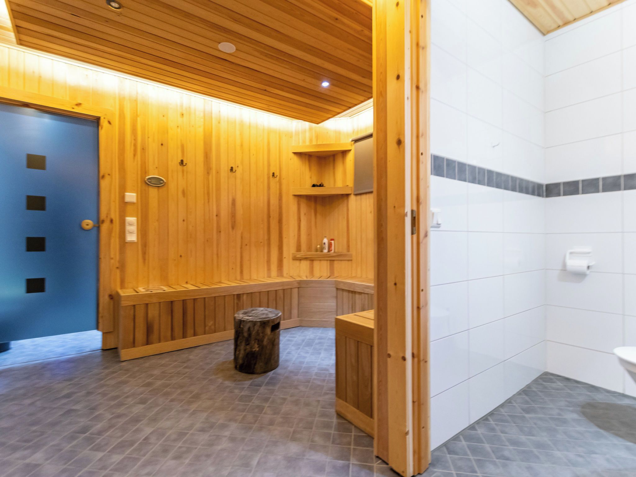 Un cuarto de sauna con bancos de madera, un taburete de madera y una pared de azulejos blancos.