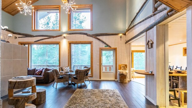 Interior acogedor de una cabaña con comedor, chimenea y ventanas amplias que ofrecen vista al bosque.