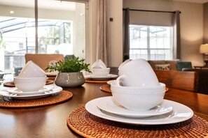 A dining table with white bowls and plates, a woven placemat, a potted plant, and a view of the outdoors through large windows.