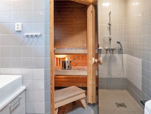 Una sauna pequeña y cerrada con un banco de madera y una olla de metal, junto a un área de ducha con un cabezal de ducha manual y un grifo empotrado en la pared.