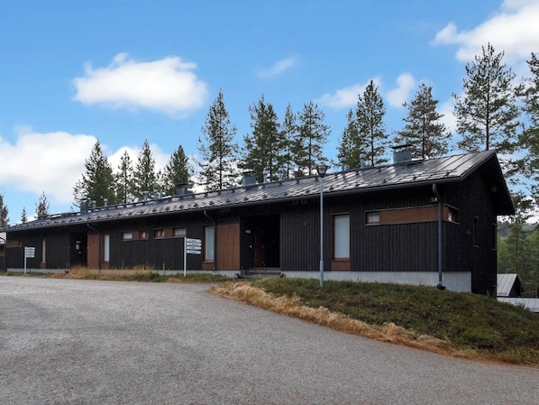 A long, single-story building with a dark exterior and a sloped roof, surrounded by a gravel driveway and dense trees.