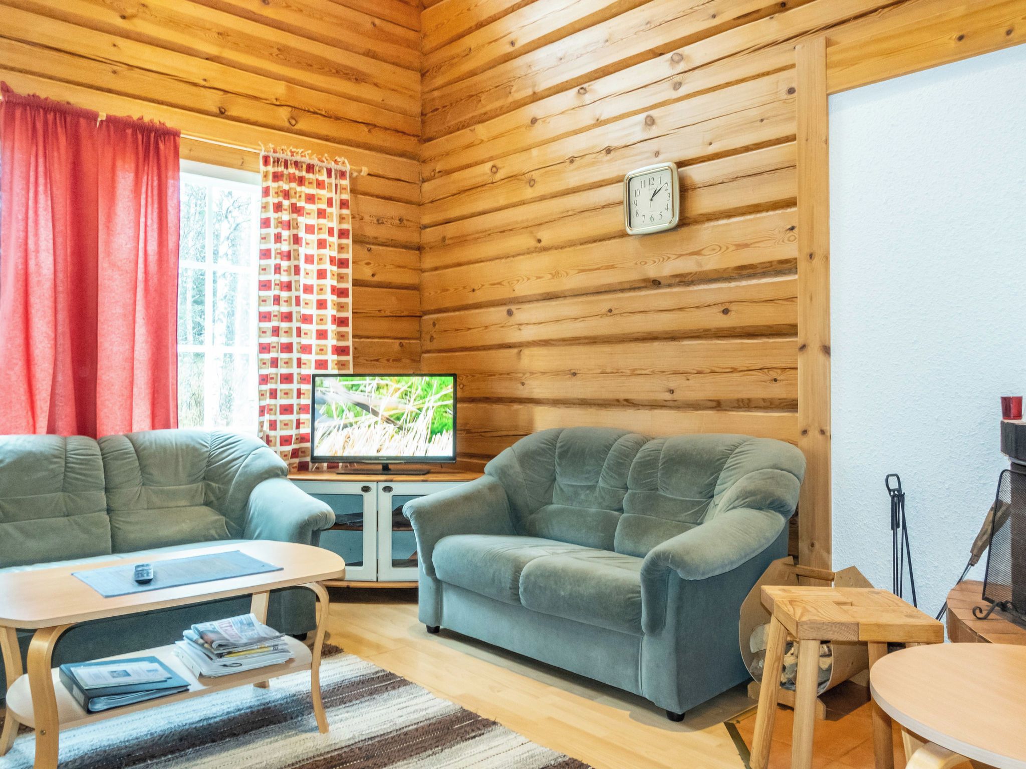 Ein gemütliches Blockhaus-Interieur mit Holzwänden, einem Fernseher, zwei Sesseln, einem kleinen Tisch und einem Fenster mit Vorhängen.