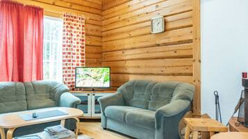 Ein gemütliches Blockhaus-Interieur mit Holzwänden, einem Fernseher, zwei Sesseln, einem kleinen Tisch und einem Fenster mit Vorhängen.