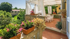 Una veranda coperta all'aperto con tavolo e sedie, circondata da piante in vaso e vista su un edificio sullo sfondo.