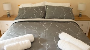 A neatly made bed with grey bedding and white pillows, two bedside tables with lamps, and two rolled towels on the bed.