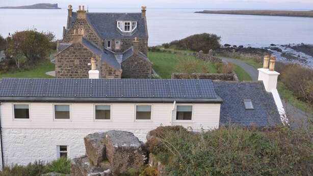A stone house with a white facade and a grey roof, overlooking a body of water.