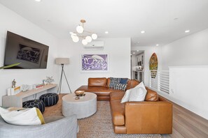A modern living room with a brown leather sofa, a round coffee table, and a grey armchair. There is a flat-screen TV mounted on the wall, a floor lamp, and artwork above the sofa.