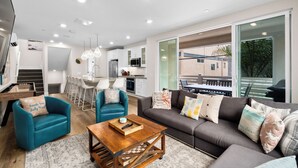 Un salon moderne avec un canapé, des fauteuils, une table basse et un coin repas comprenant des chaises, et une cuisine en arrière-plan.