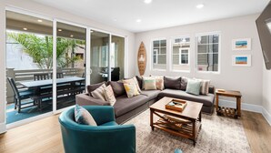 Un salon moderne avec un canapé d’angle, un fauteuil turquoise et une table basse en bois. Une porte vitrée donne accès à un patio extérieur aménagé avec une table et des chaises.