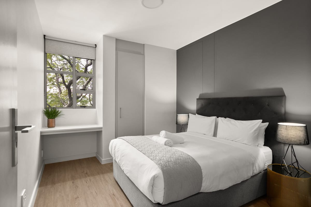 A modern bedroom with a large bed, a window with a view, a bedside table, and a lamp.