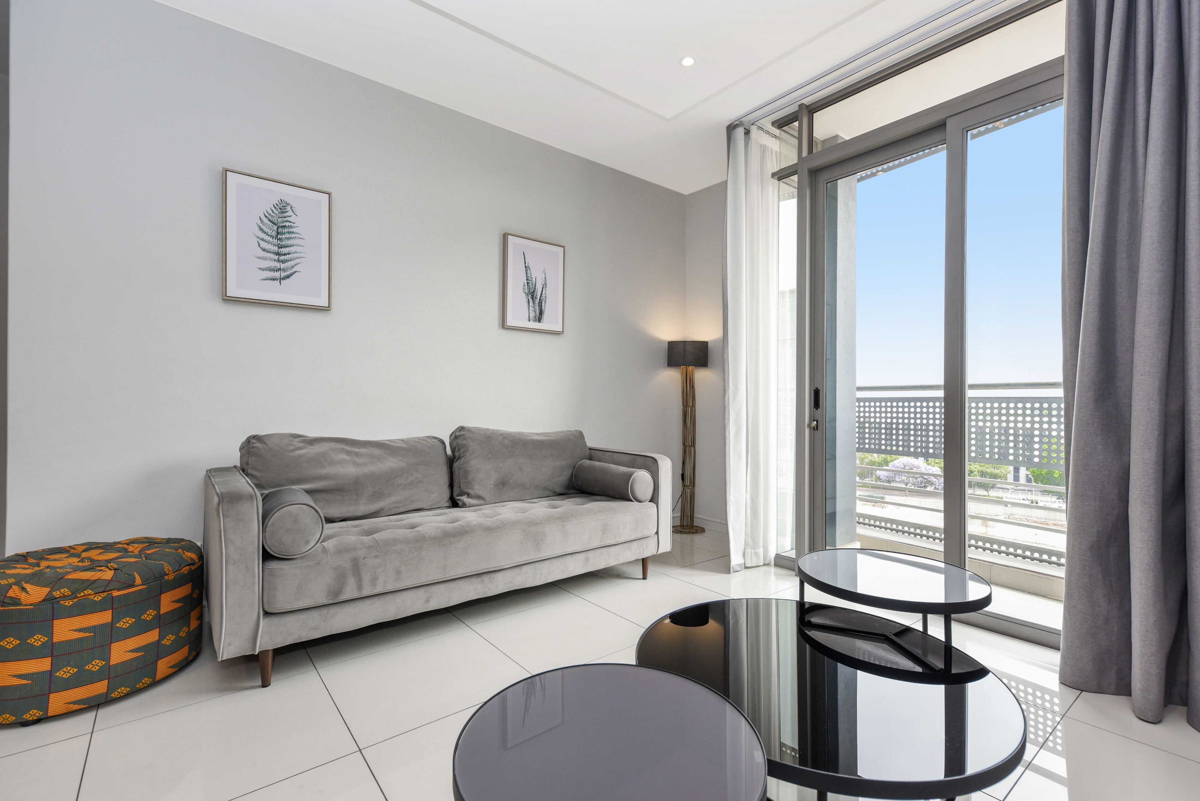 Un salon moderne avec un canapé gris, une table ronde en verre et un pouf à motifs. Deux œuvres d’art encadrées ornent le mur, et une lampe sur pied est placée dans un coin.