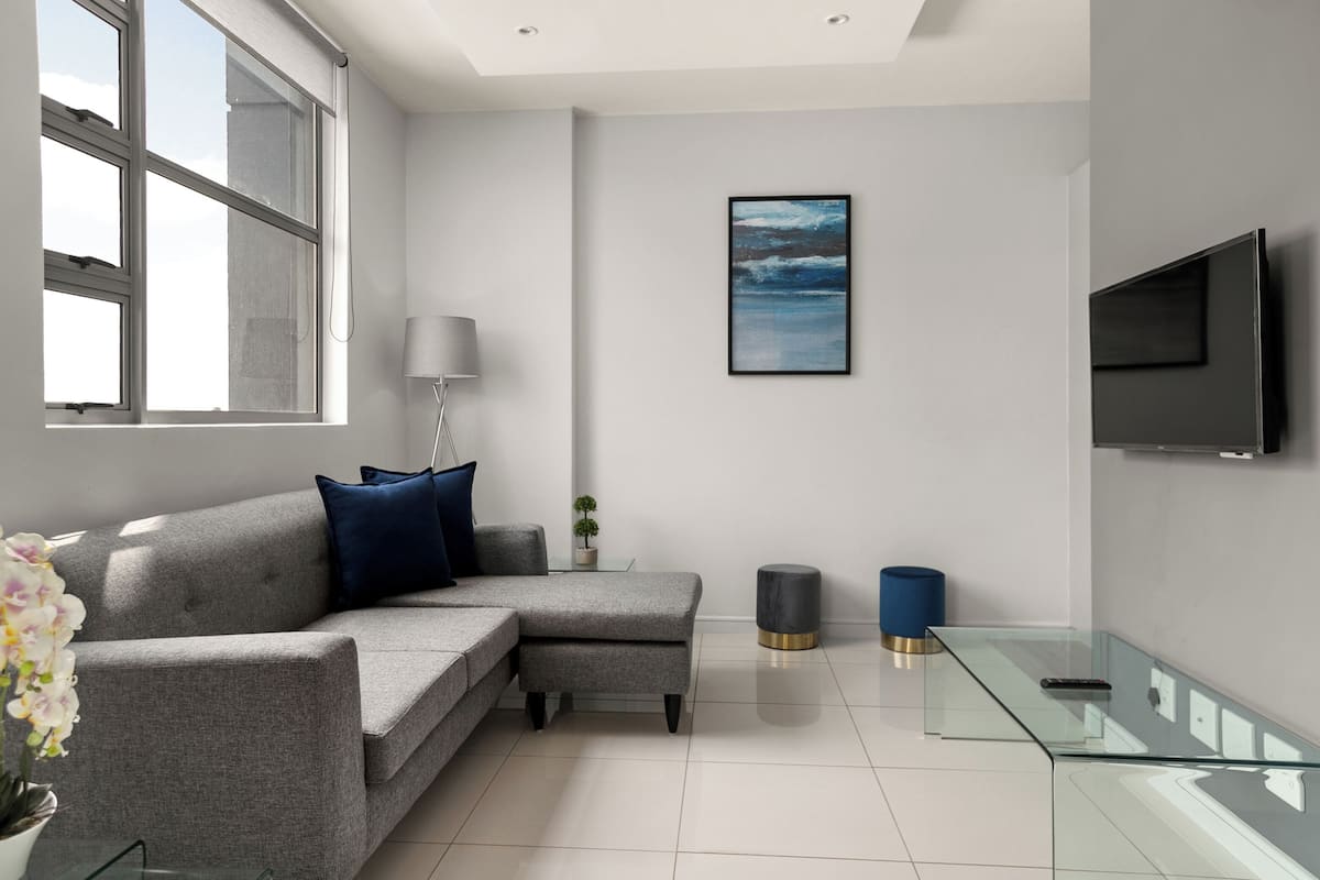 A modern living room with a grey sectional sofa, a glass coffee table, a mounted TV, and a framed artwork on the wall.