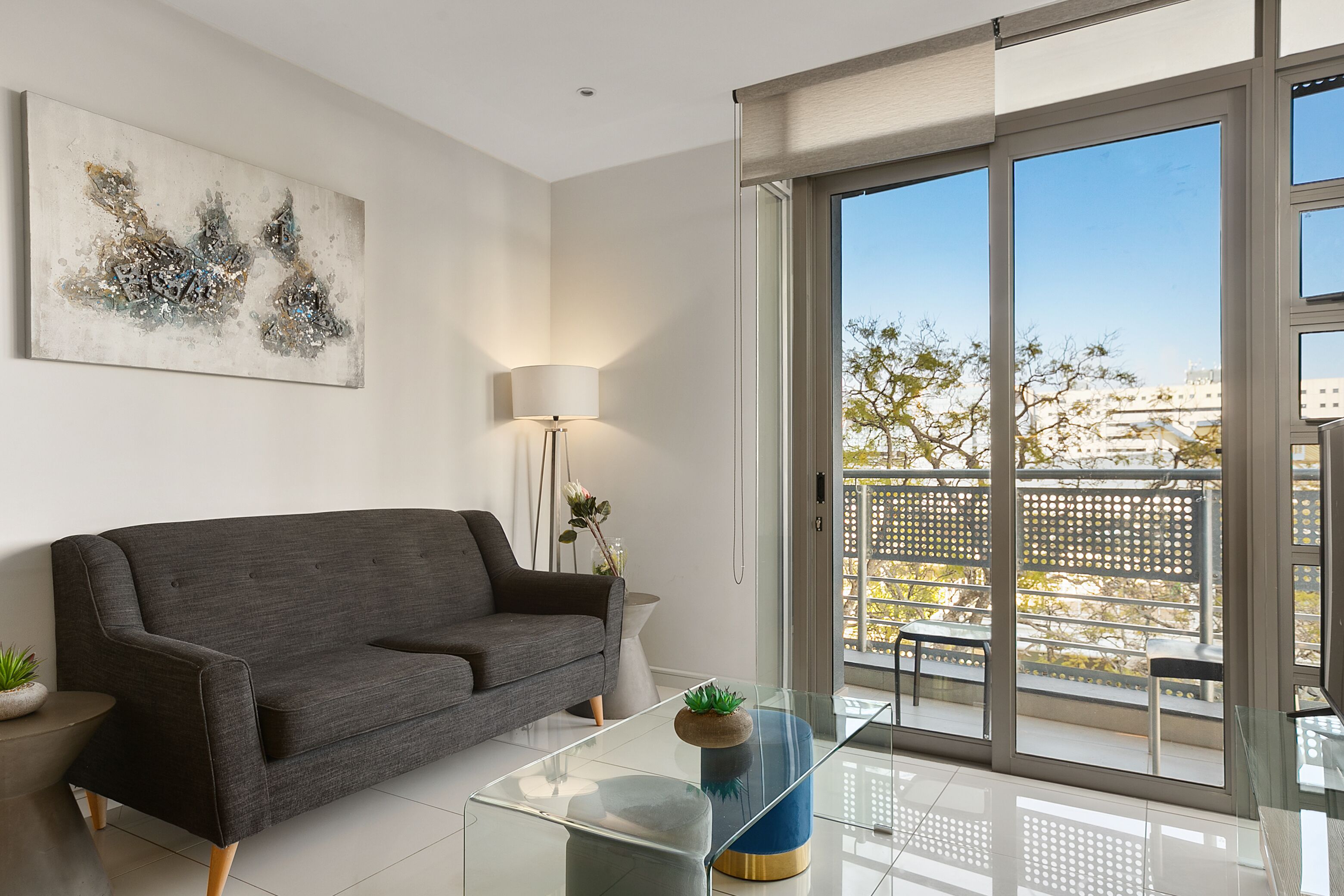 Een moderne woonkamer met een grijze bank, een glazen salontafel en een groot raam met uitzicht op de stad.