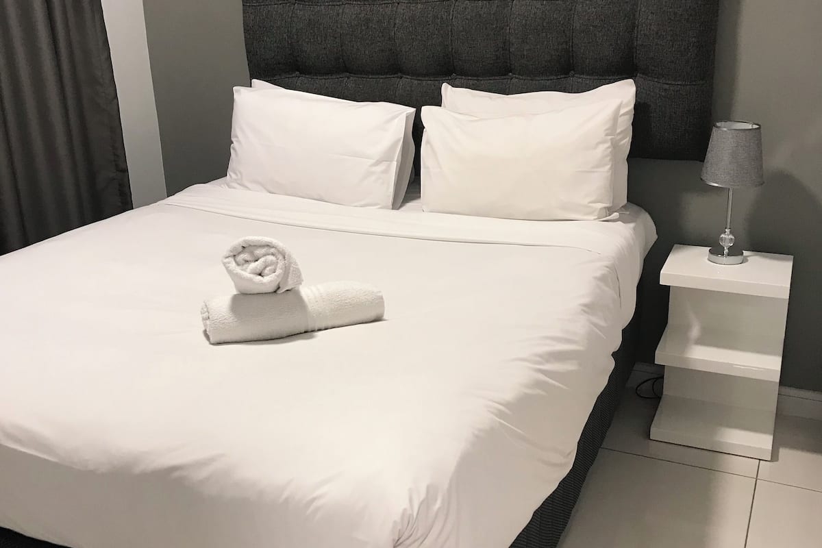 A neatly made bed with white linens and a rolled towel, a dark upholstered headboard, and a bedside table with a lamp.