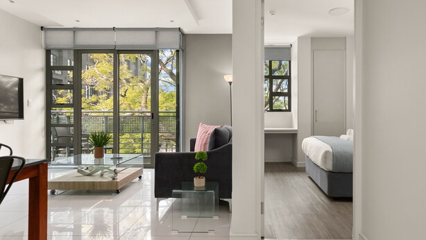 Een moderne woonkamer met een glazen salontafel, een bank en een bed in de aangrenzende kamer.