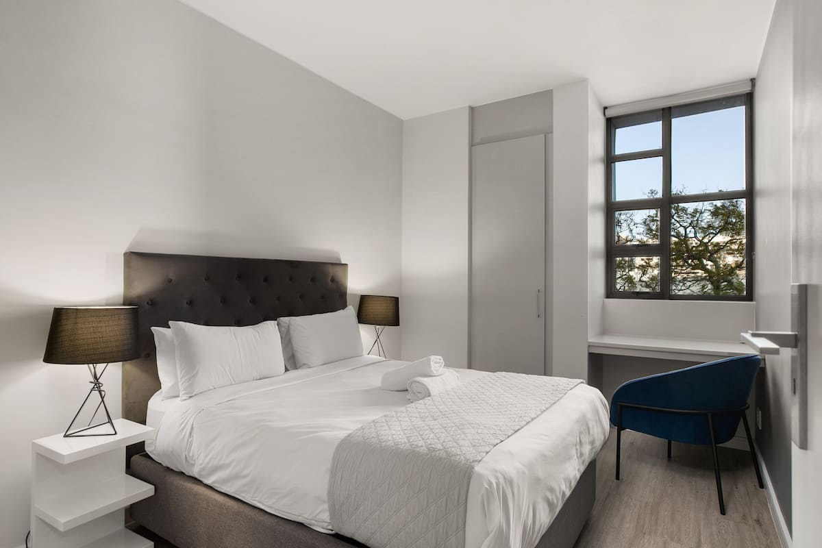 A modern bedroom with a large bed, two bedside tables with lamps, a desk, and a chair.