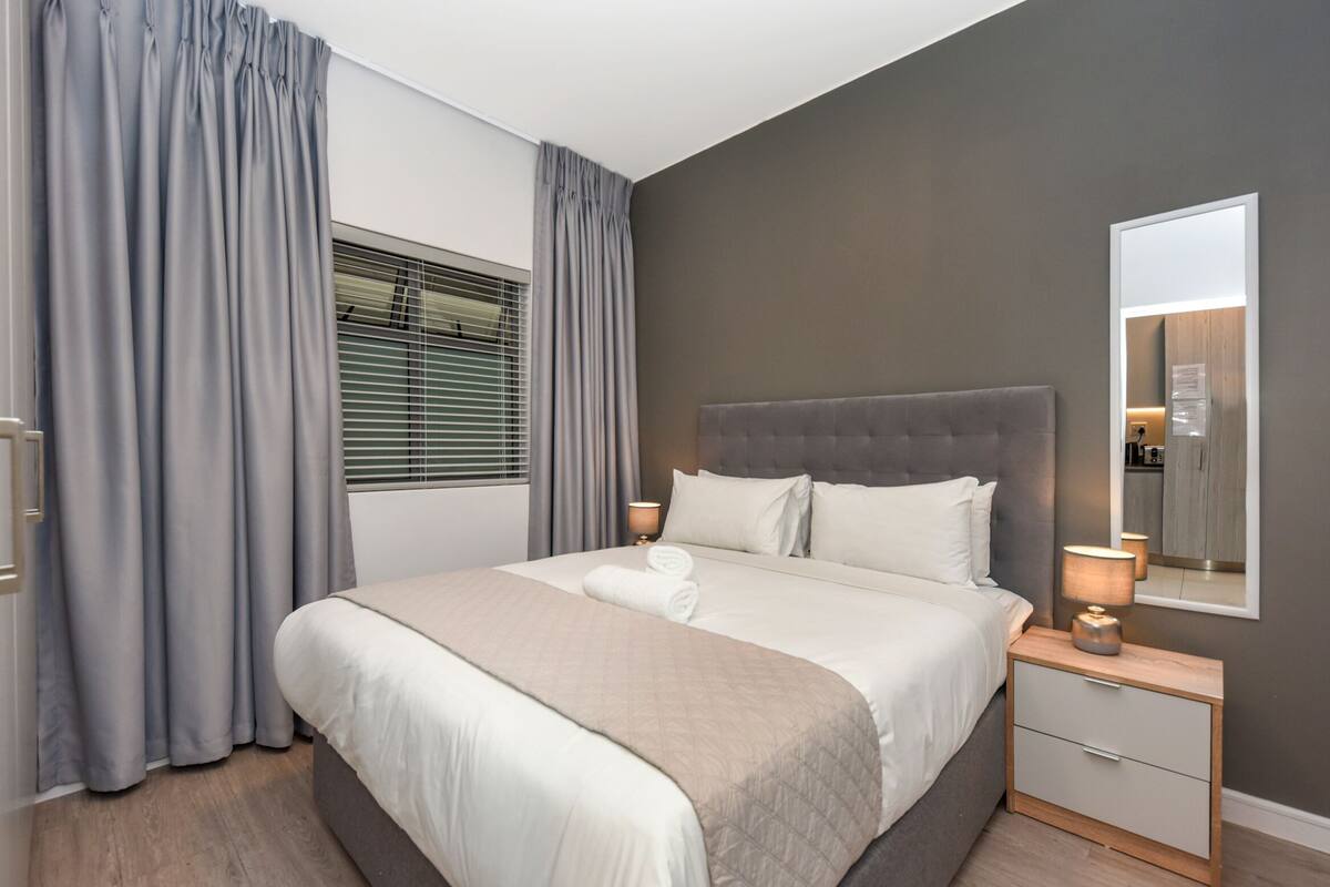 A modern bedroom with a large bed, grey curtains, a bedside lamp, and a mirror on the wall.