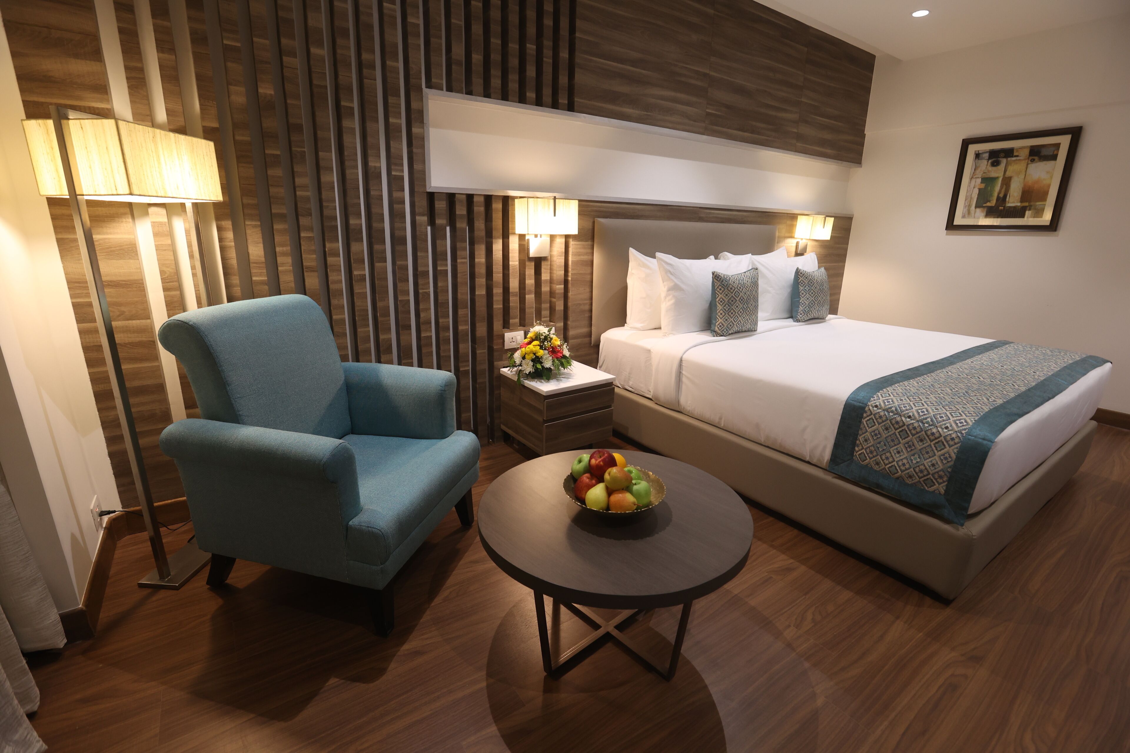 Une chambre d’hôtel avec un lit, un fauteuil bleu, une petite table avec des fruits et une lampe.
