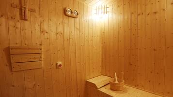 En bastu med träpanel, en träbänk, en väggmonterad värmare och en trähink med en öskar.