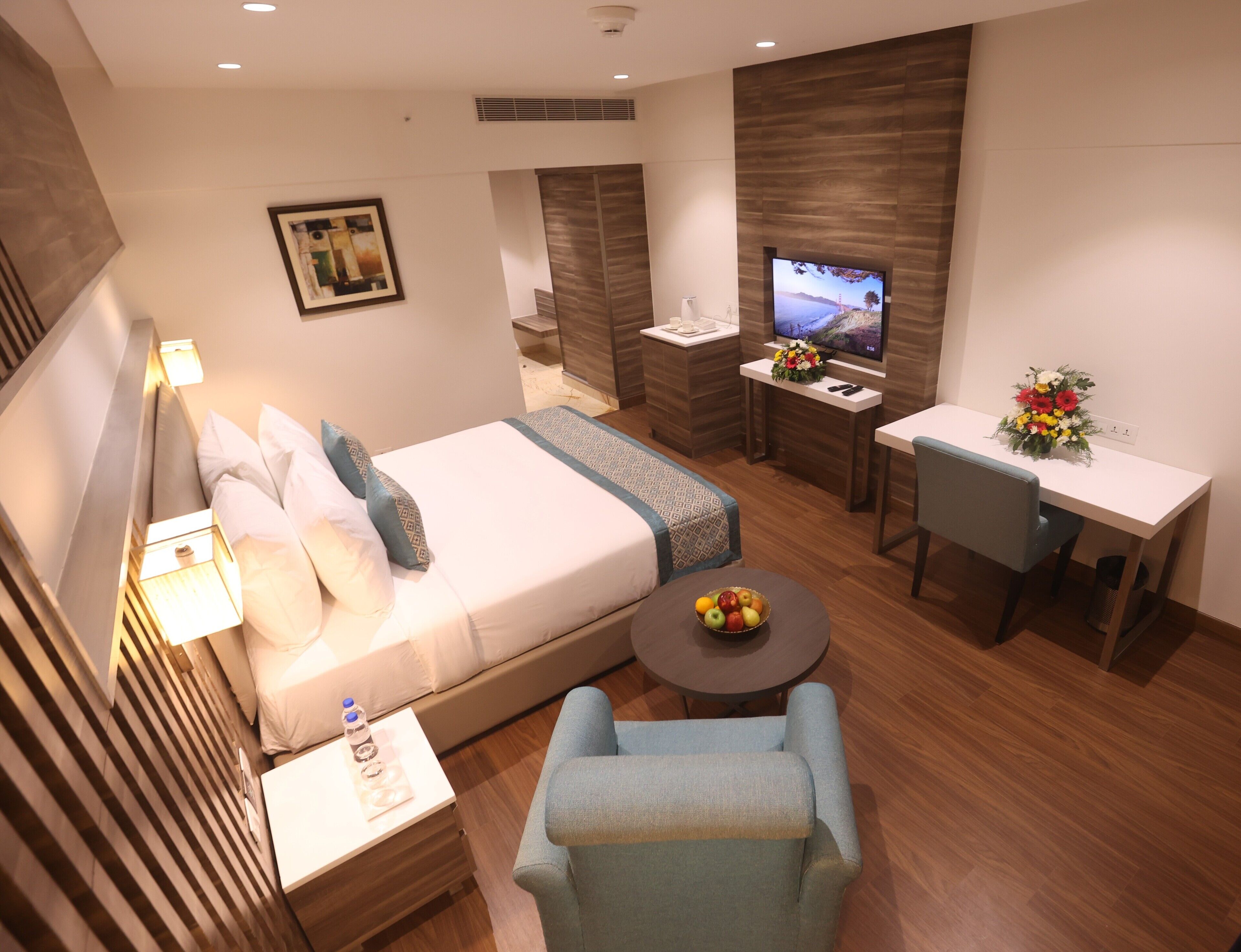 A modern hotel room with a bed, a desk, a TV, and a seating area.