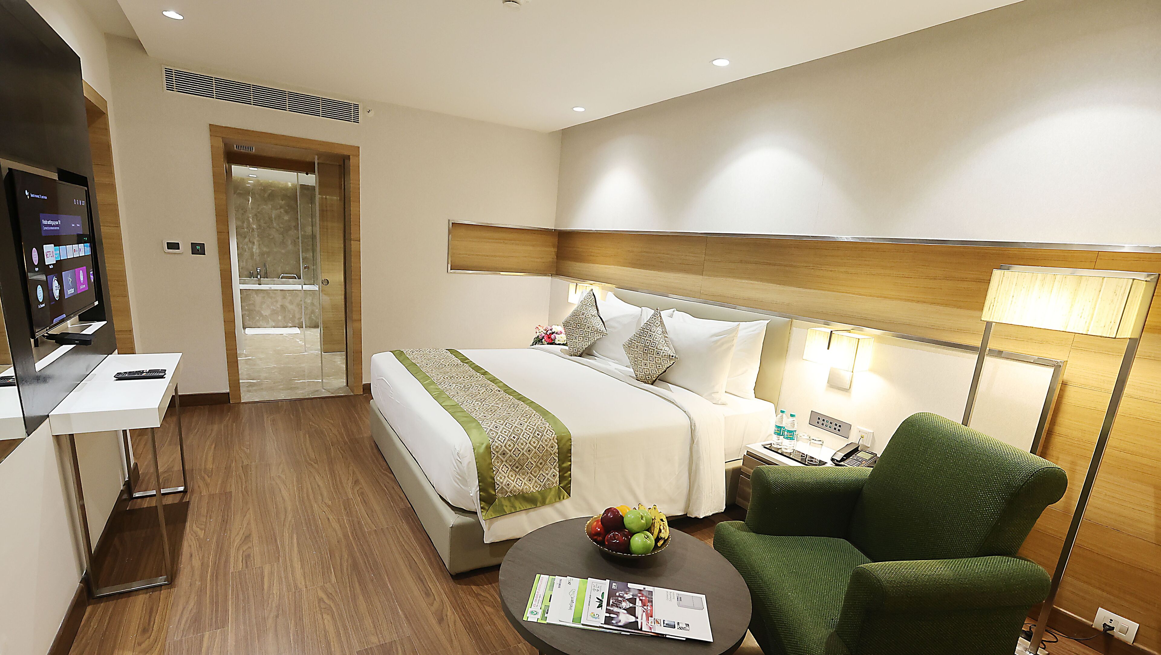 Habitación de hotel con cama, escritorio con televisor, silla, mesa con fruta y baño visible a través de una puerta de cristal.