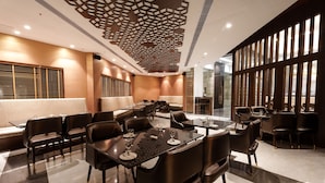 A modern dining area with black leather chairs and tables, a geometric ceiling design, and large windows providing natural light. - Pharos Hotels (Chennai)