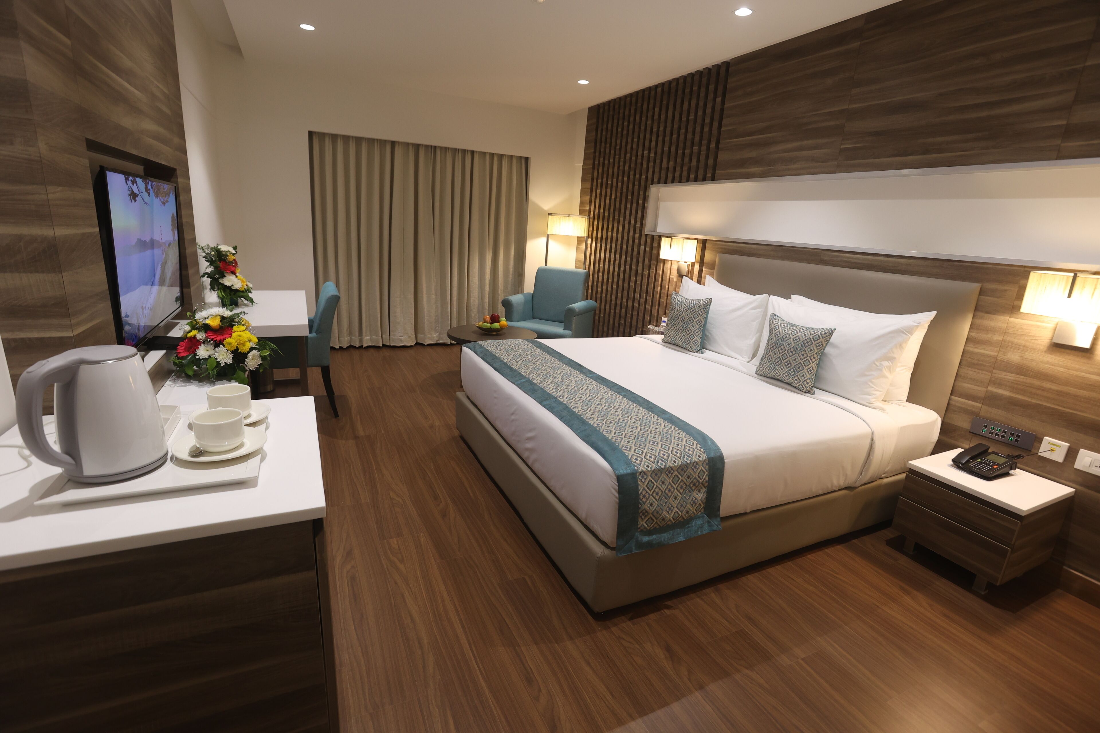 A modern hotel room with a large bed, a desk with a kettle, a chair, a TV, and a decorative plant.