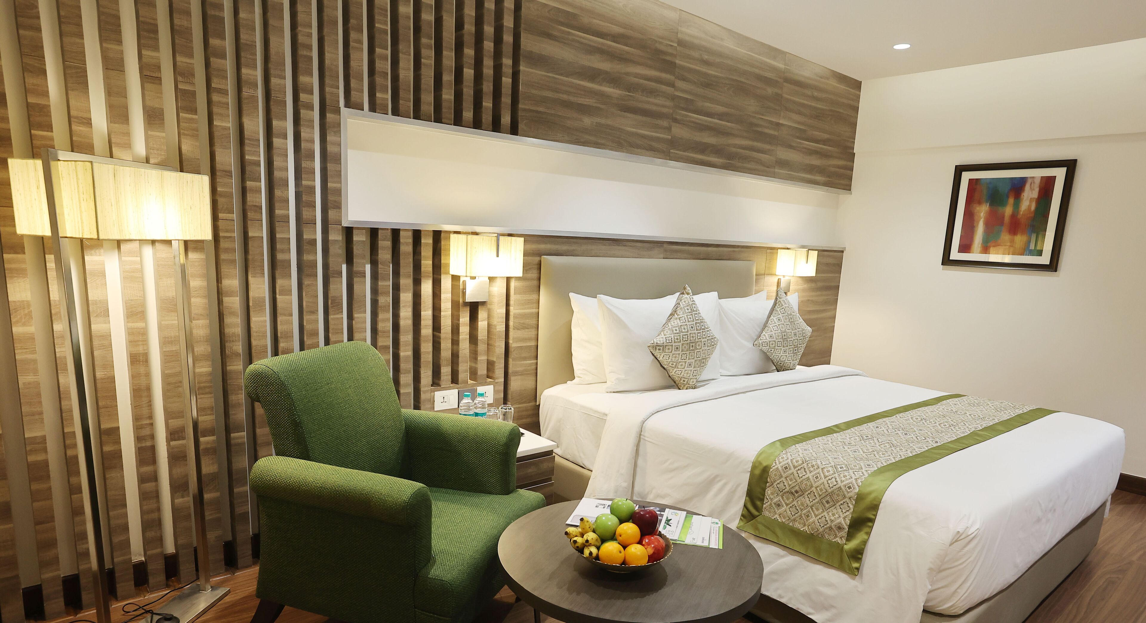 Una habitación de hotel con una cama, un sillón verde, una mesita con fruta y una lámpara de pared.