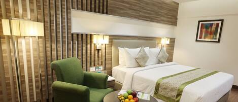 A hotel room with a bed, a green armchair, a small table with fruit, and a wall-mounted lamp.