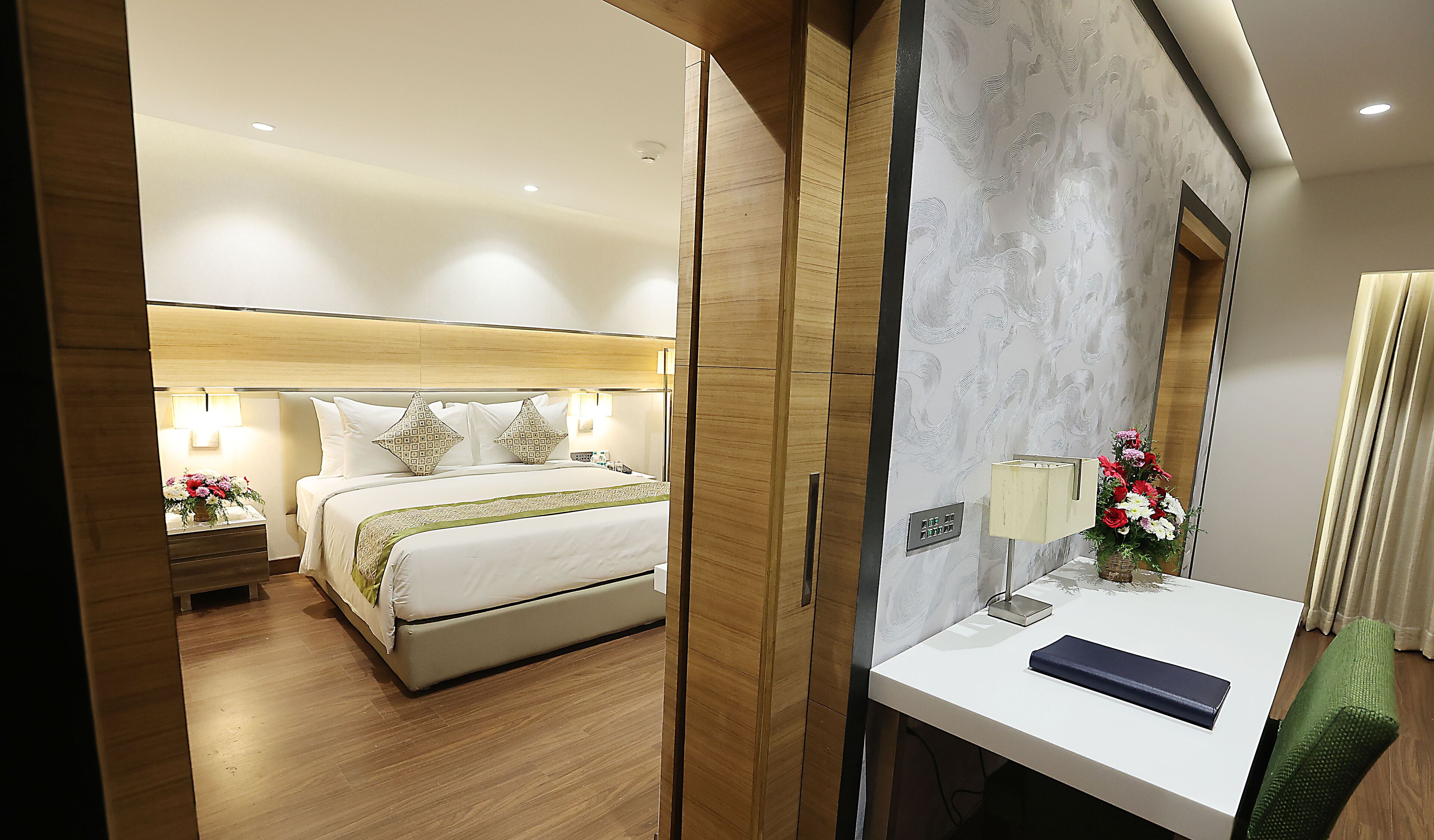 A hotel room with a bed, bedside tables, a desk, a chair, a mirror, and a flower arrangement.