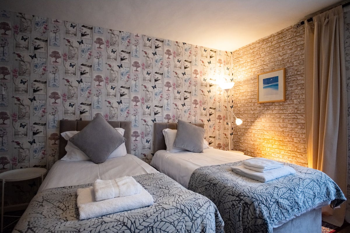 A hotel room with two beds, floral wallpaper, a brick wall, a night sky picture, and a bedside lamp.