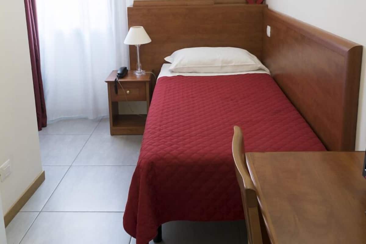 A hotel room with a wooden bed, a nightstand, a table, a chair, a painting on the wall, and a window with curtains.