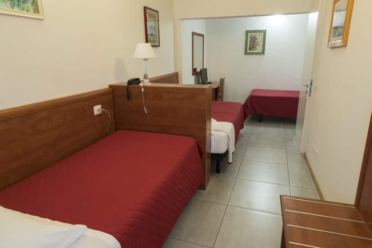 A hotel room with two single beds, a desk with a chair, a small table with a red tablecloth, and a mirror on the wall.