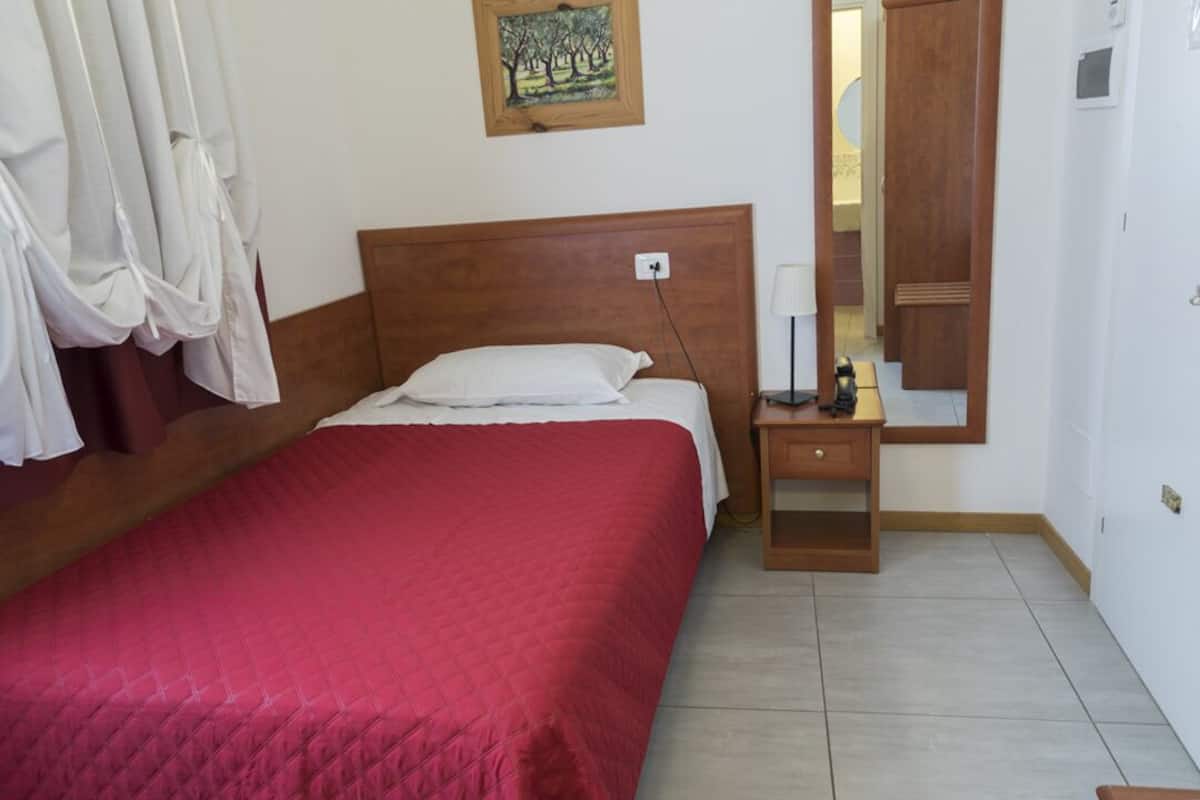 A bedroom with a wooden headboard, a red bedspread, a bedside table with a lamp, and a mirror.