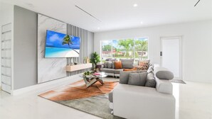 Ein modernes Wohnzimmer mit großem Fernseher, einer Couch, einem Couchtisch und Blick auf eine tropische Landschaft durch das Fenster.