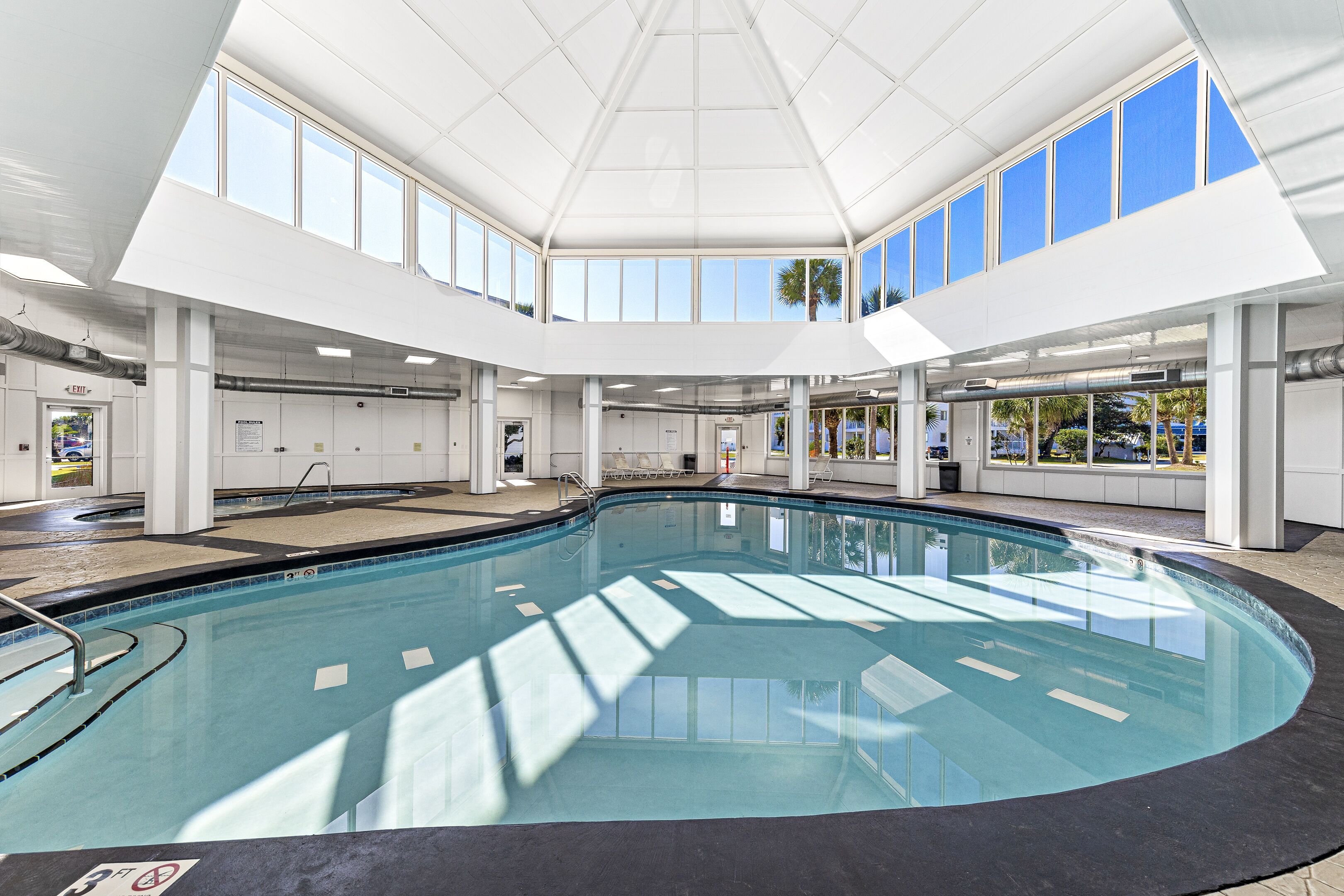 A large indoor swimming pool with clear glass walls, multiple exits, and a high ceiling.
