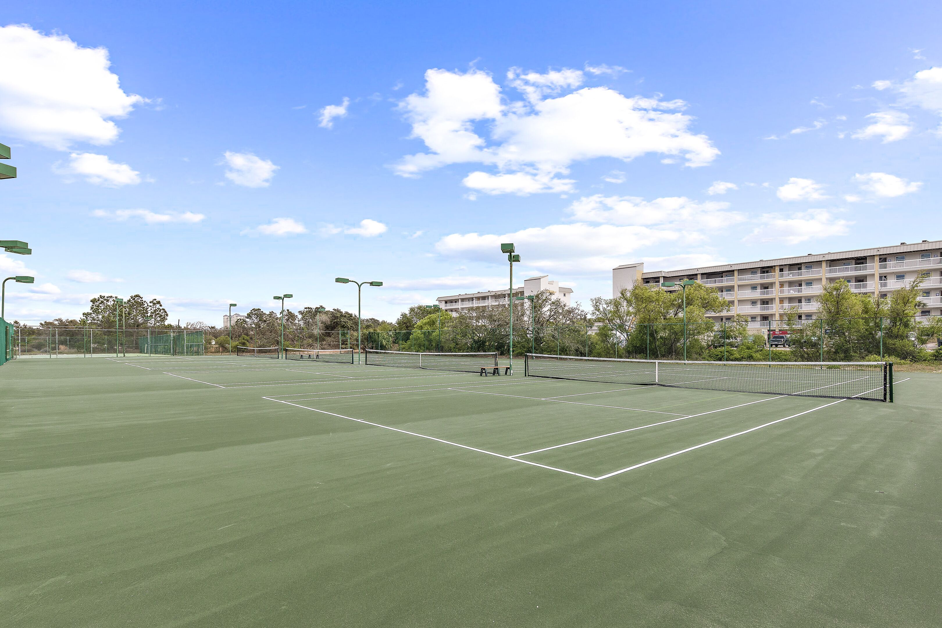 Una cancha de tenis al aire libre con una red, rodeada de árboles y un edificio al fondo.