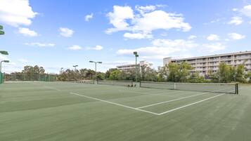 Una cancha de tenis al aire libre con una red, rodeada de árboles y un edificio al fondo.