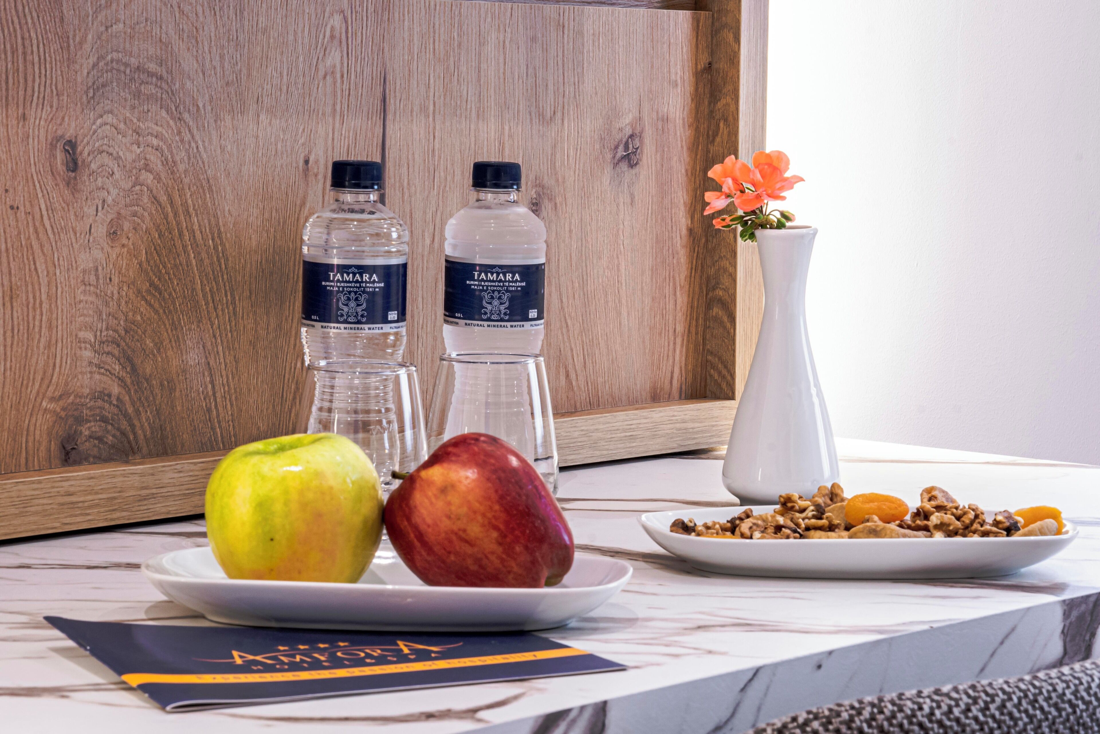 Ein Tisch mit zwei Wasserflaschen, einem Teller mit einem Apfel und einer roten Frucht, einem Teller mit Nüssen und einer Vase mit Blumen.