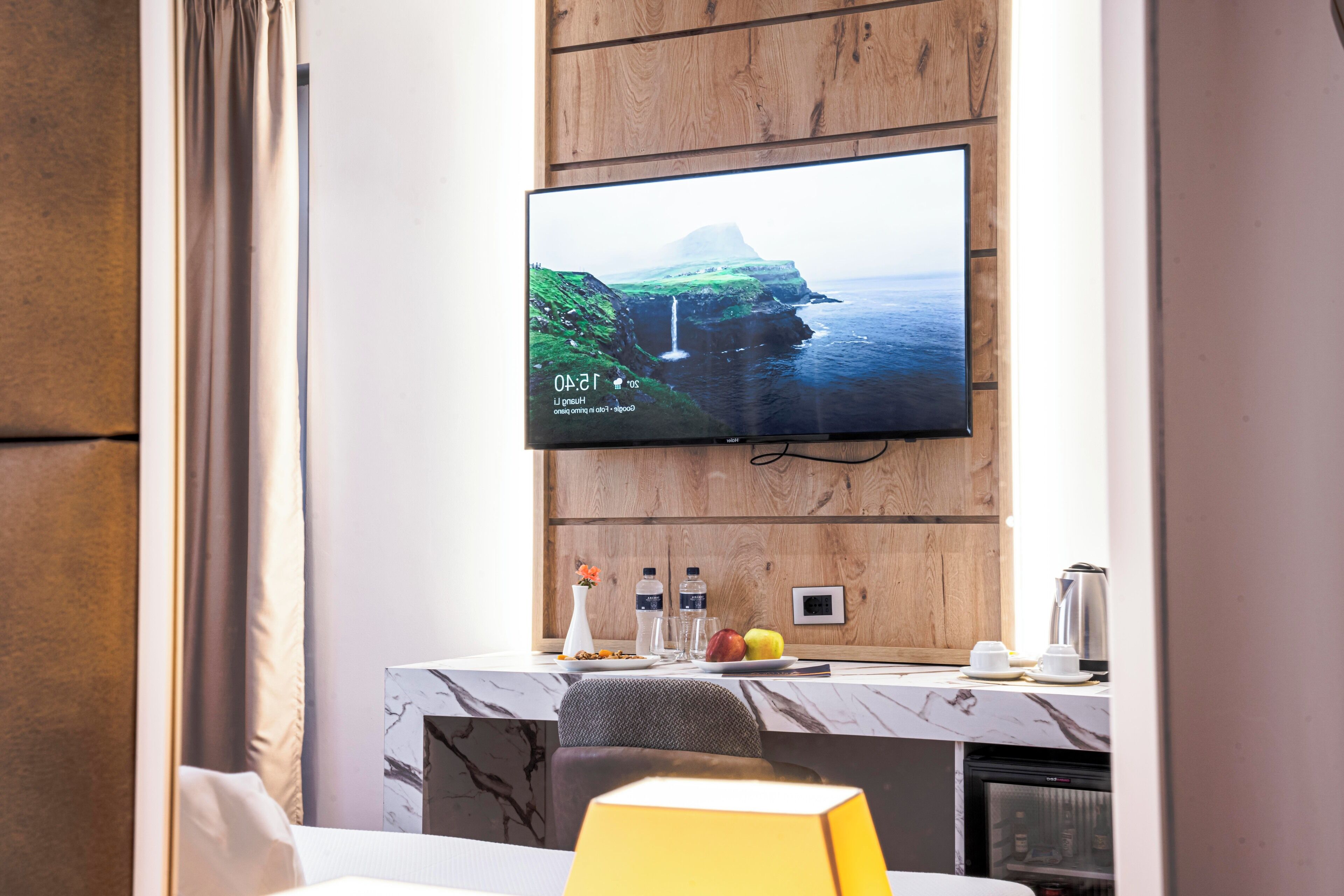 Habitación de hotel con televisor de pantalla plana montado en una pared de madera, encimera de mármol, un refrigerador pequeño y una zona de estar con una silla y una mesa.