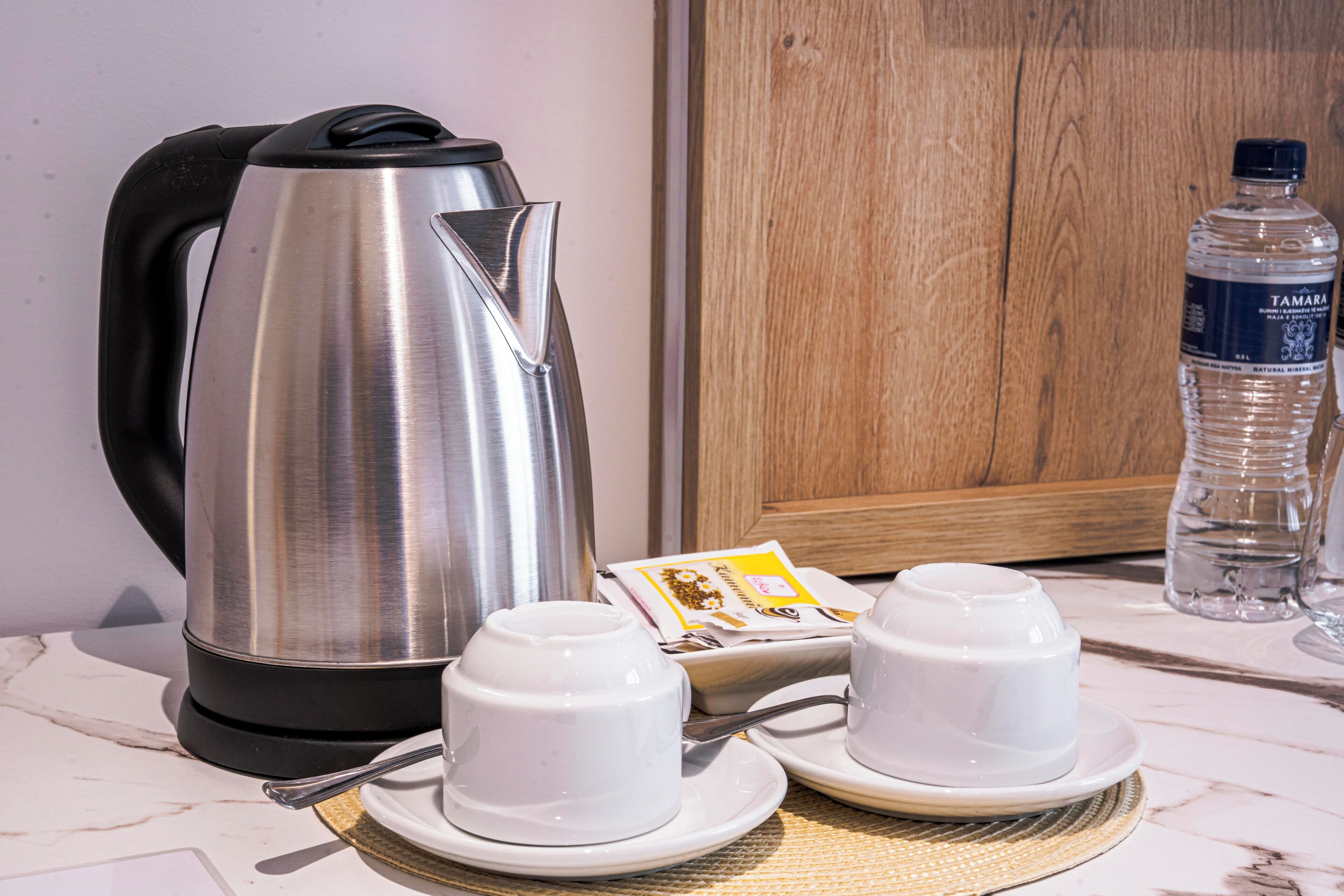 A stainless steel kettle, two white teacups with saucers, a water bottle, and a wooden cabinet in a kitchen setting.